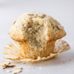 A partially eaten poppy seed muffin with a sugar-dusted top and almond slices, sitting on an opened muffin wrapper against a light background.