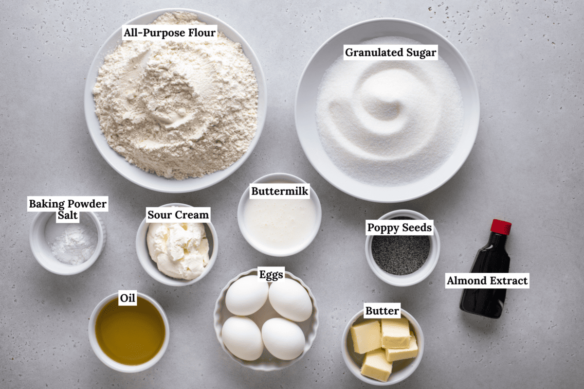 Top-down view of labeled baking ingredients in bowls: all-purpose flour, granulated sugar, baking powder, salt, sour cream, buttermilk, poppy seeds, eggs, almond extract, butter, and oil on a light surface.
