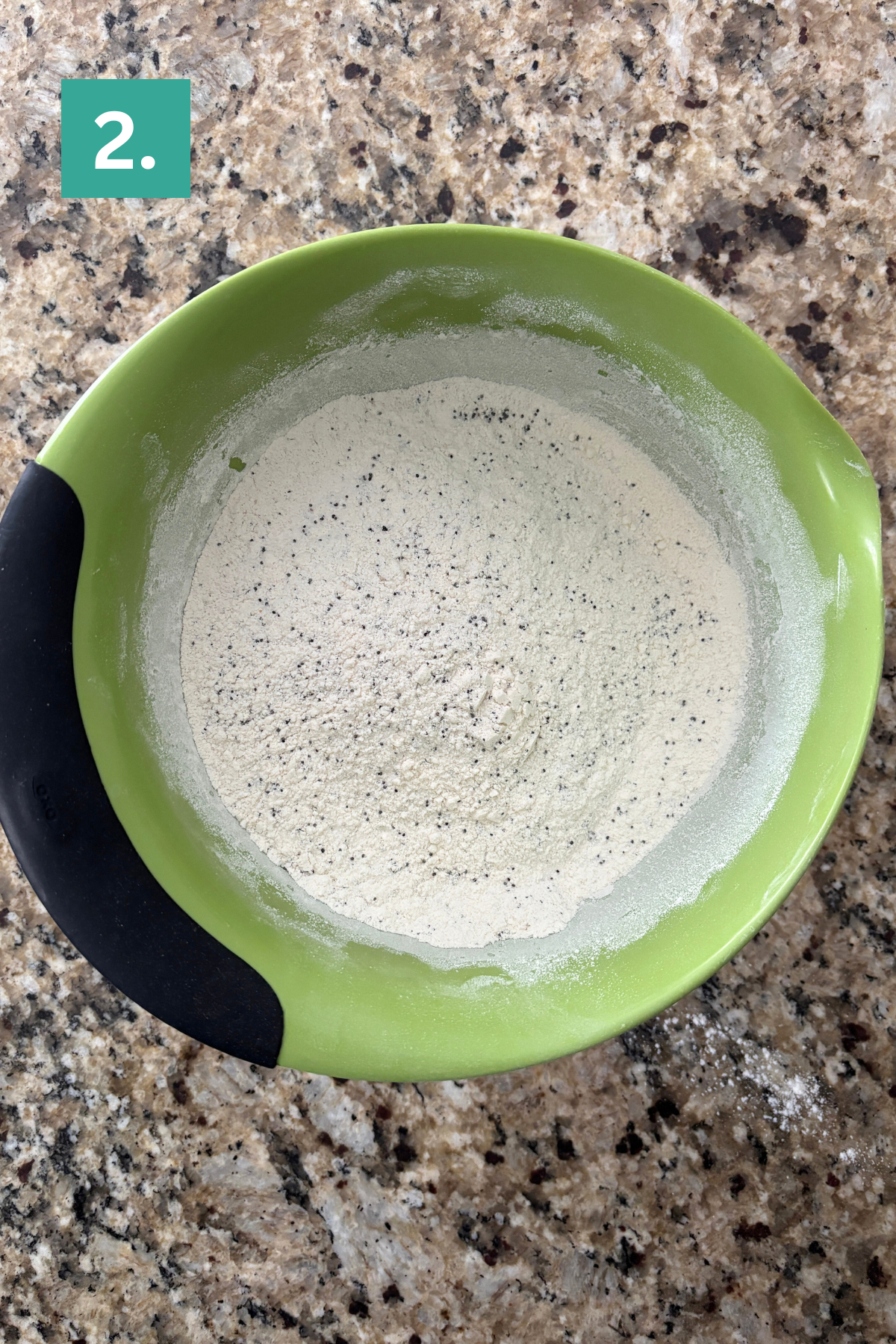A green mixing bowl with black grip holds a mixture of flour and specks of seasoning on a granite countertop. A green box with the number 2 is in the top left corner.