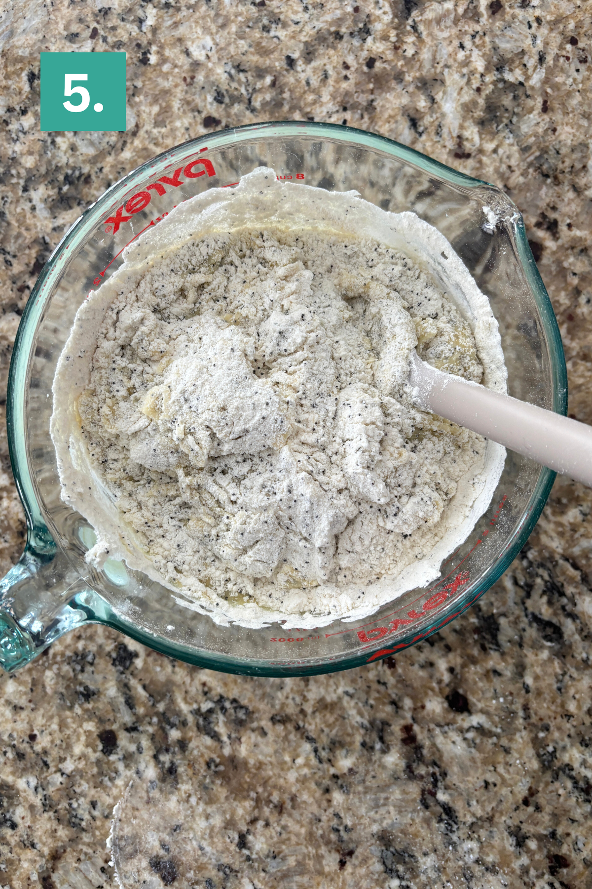 A glass measuring cup filled with flour mixture and a white spatula sits on a granite countertop. A green box with the number 5 is in the upper left corner of the image.