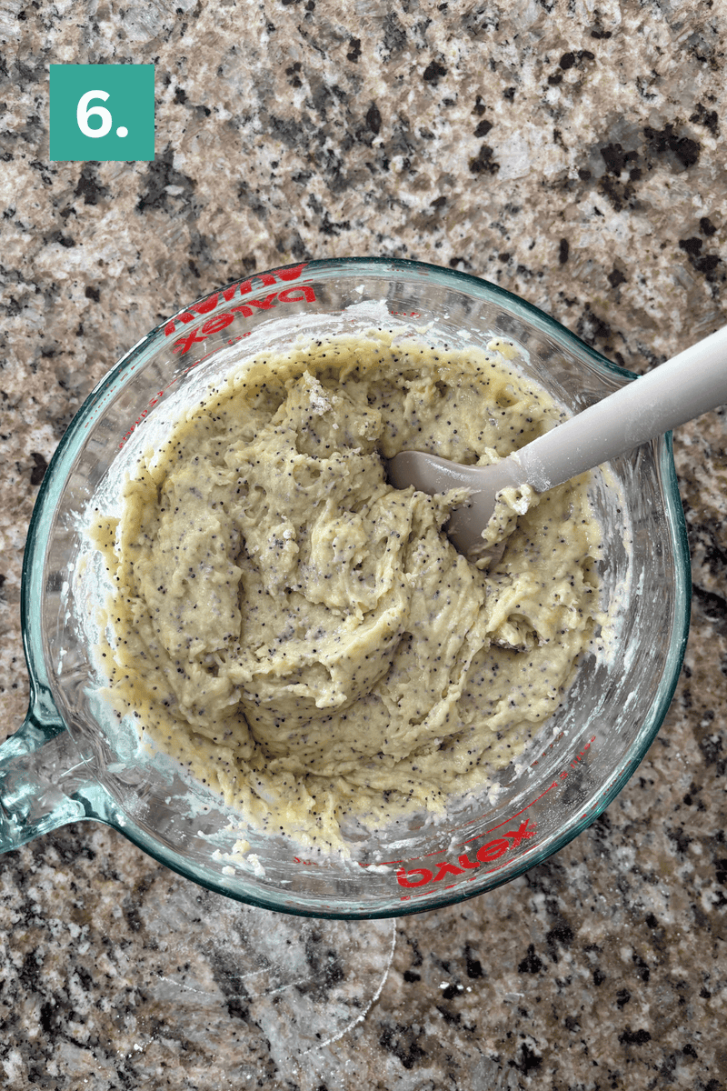 A glass measuring cup filled with a thick, pale batter mixed with poppy seeds is being stirred with a gray spatula. The cup sits on a speckled kitchen counter. A green box with the number 6 is in the top left corner.