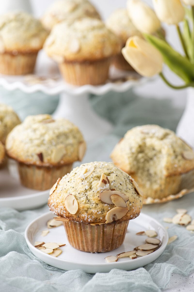A poppy seed muffin topped with sliced almonds sits on a small white plate. More muffins and a bouquet of yellow tulips are blurred in the background.
