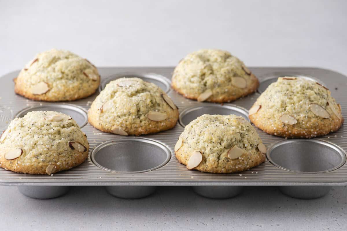 A muffin tin with six baked almond muffins, each topped with sliced almonds and sugar, sits on a light gray surface. Two of the muffin cups are empty.