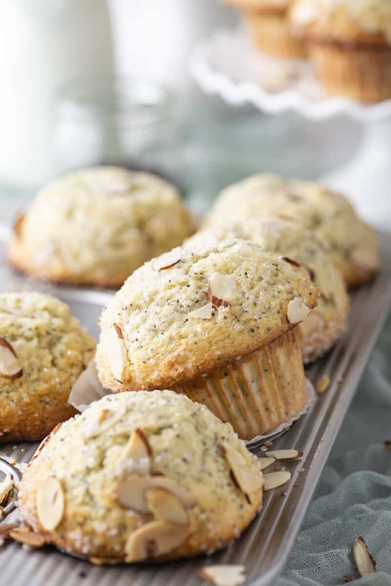 Freshly baked almond poppy seed muffins are arranged on a metal cooling rack, topped with sliced almonds and coarse sugar. More muffins and a glass jar appear softly blurred in the background.