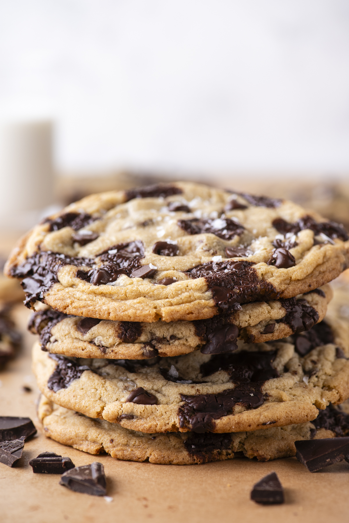 A stack of four large, chewy chocolate chip cookies sits on a brown parchment surface, with melted chocolate chunks visible and a few scattered chocolate pieces around. The background is softly blurred.