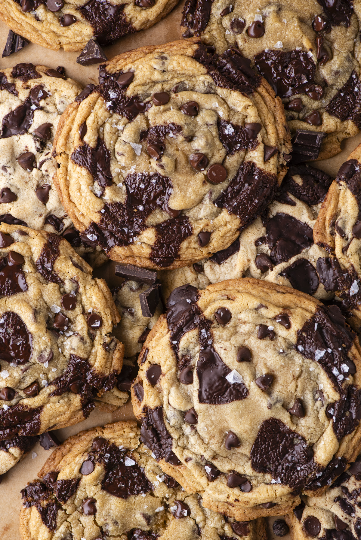 A close-up of several chocolate chip cookies with large chunks of melted chocolate and a sprinkle of sea salt on top, overlapping each other on a light brown surface.