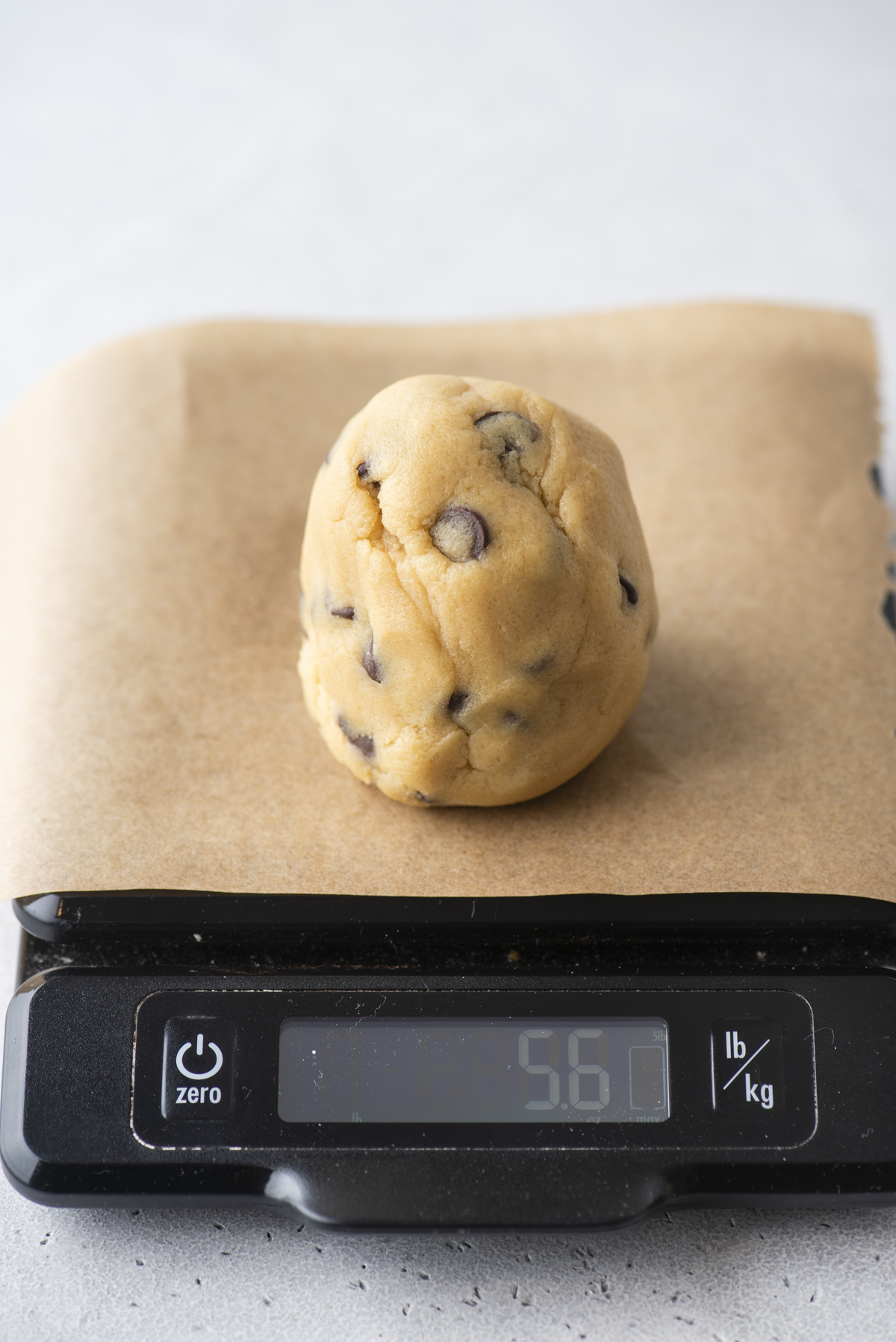 A ball of chocolate chip cookie dough sits on parchment paper atop a digital kitchen scale, which displays a weight of 5.6 ounces.