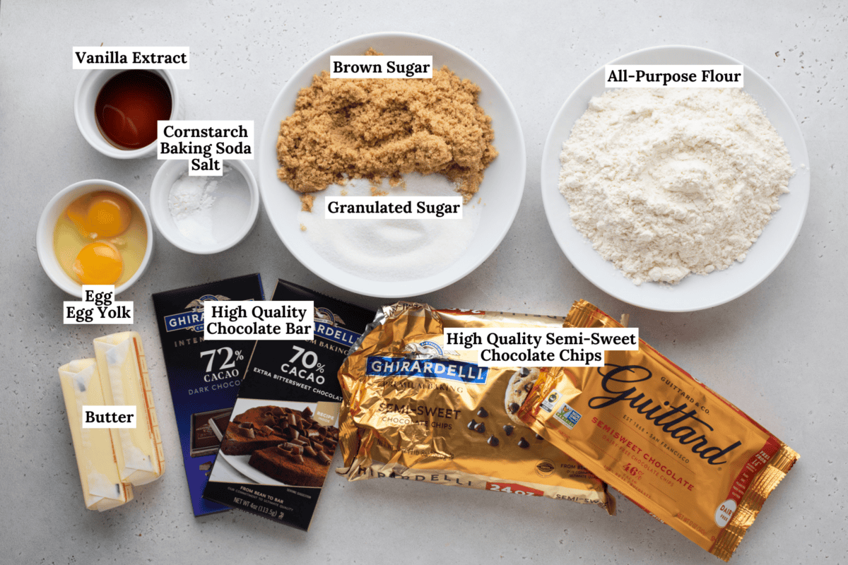 Overhead view of labeled chocolate chip cookie ingredients: vanilla extract, brown sugar, all-purpose flour, cornstarch, baking soda, salt, granulated sugar, eggs, butter, chocolate bars, and chocolate chips.