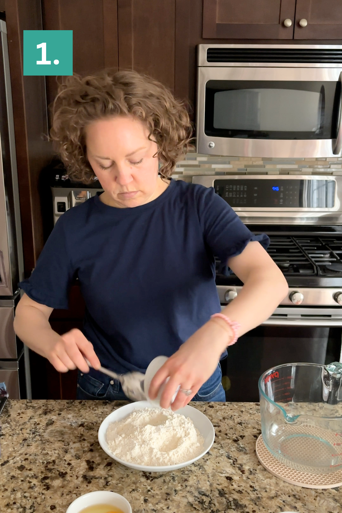 A woman in a navy t-shirt stands in a kitchen, preparing bakery style chocolate chip cookies as she pours an ingredient into a bowl of flour on the granite countertop. A microwave and oven are behind her. A green box with 1. is in the upper left corner.
