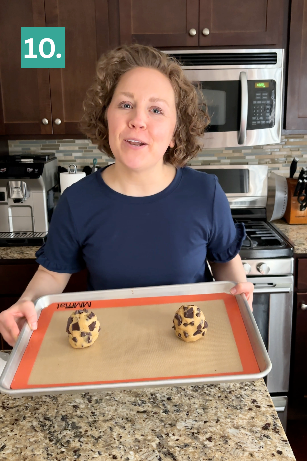 A person with short curly hair, wearing a navy blue shirt, holds a baking tray with two large balls of bakery style chocolate chip cookie dough in a kitchen. A green square with the number 10 is in the upper left corner.