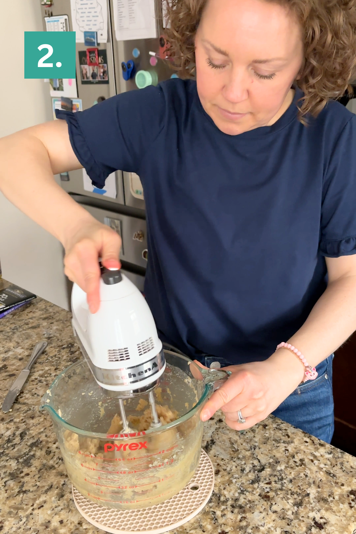 A woman mixes ingredients in a glass bowl with a hand mixer on a granite kitchen counter, preparing bakery style chocolate chip cookies. She wears a navy shirt and jeans. A green box with the number 2 is in the top left corner.