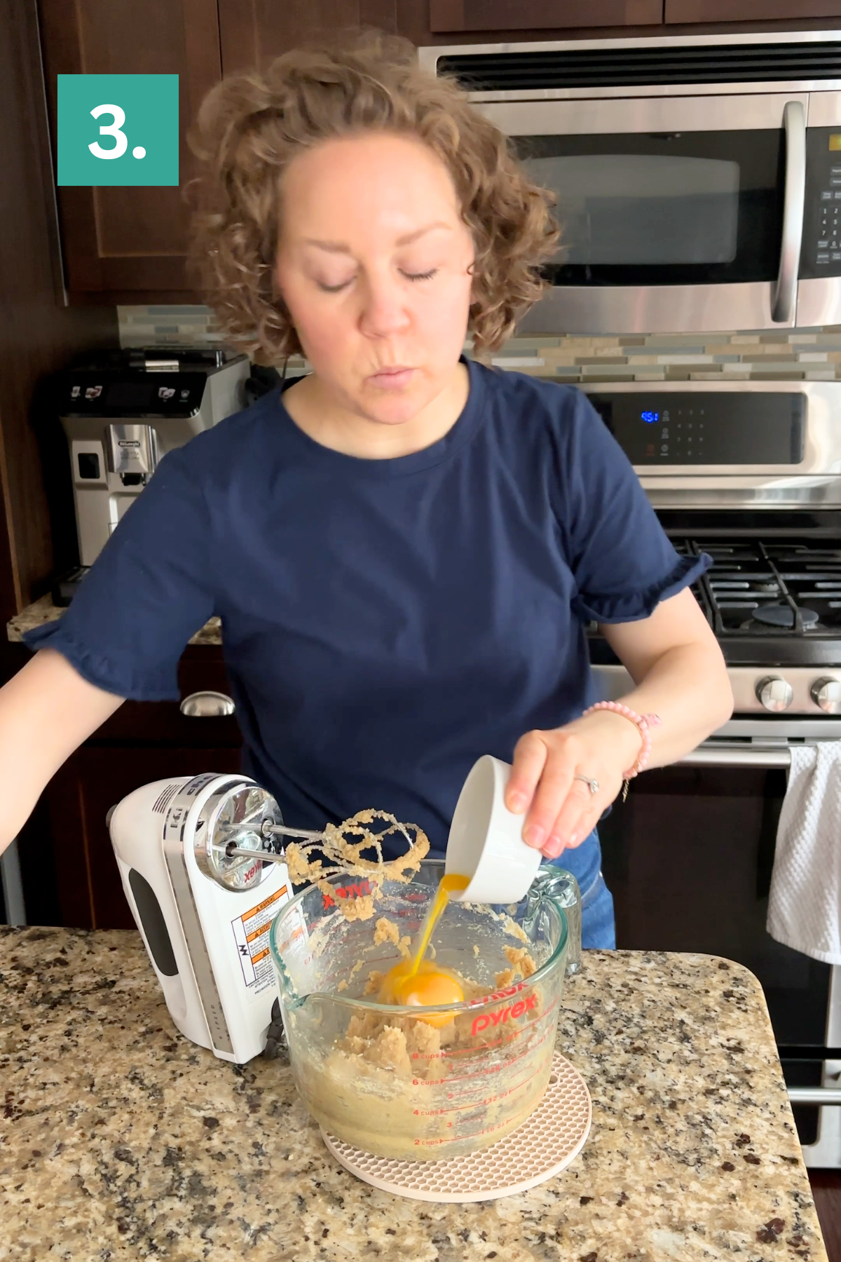 A woman in a navy blue shirt stands in a kitchen, pouring beaten eggs into a glass mixing bowl with batter for bakery style chocolate chip cookies. A hand mixer rests on the counter nearby. A green box with 3. is in the top left corner.