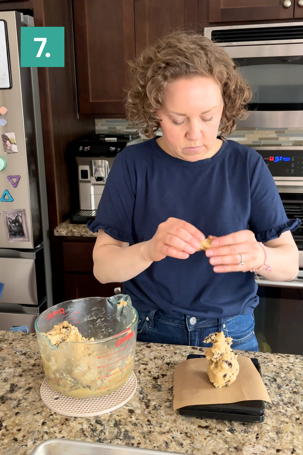 A woman stands in a kitchen shaping bakery style chocolate chip cookies with her hands. Beside her is a large measuring cup of dough, and portions rest on parchment paper. A green box with the number 7 appears at the top left.