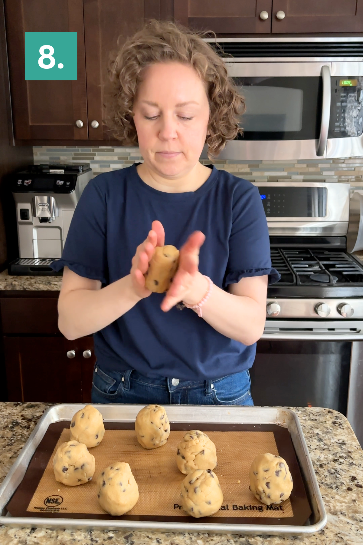 A woman in a navy shirt stands in a kitchen, shaping bakery style chocolate chip cookie dough into balls over a baking sheet with seven dough balls. A green square with the number 8 appears in the upper left corner.