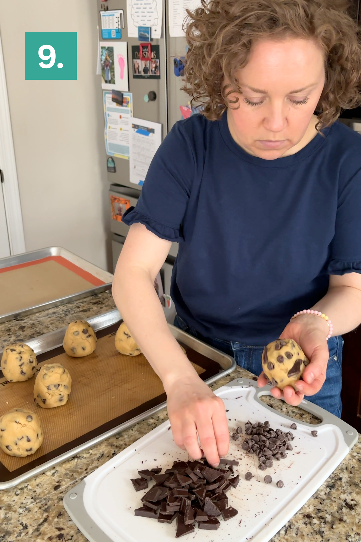 A woman in a navy shirt adds chocolate pieces to dough balls in a kitchen, creating bakery style chocolate chip cookies. Several dough balls are on a baking tray, with chopped chocolate and chips on a white cutting board. A green box with the number 9 is in the corner.