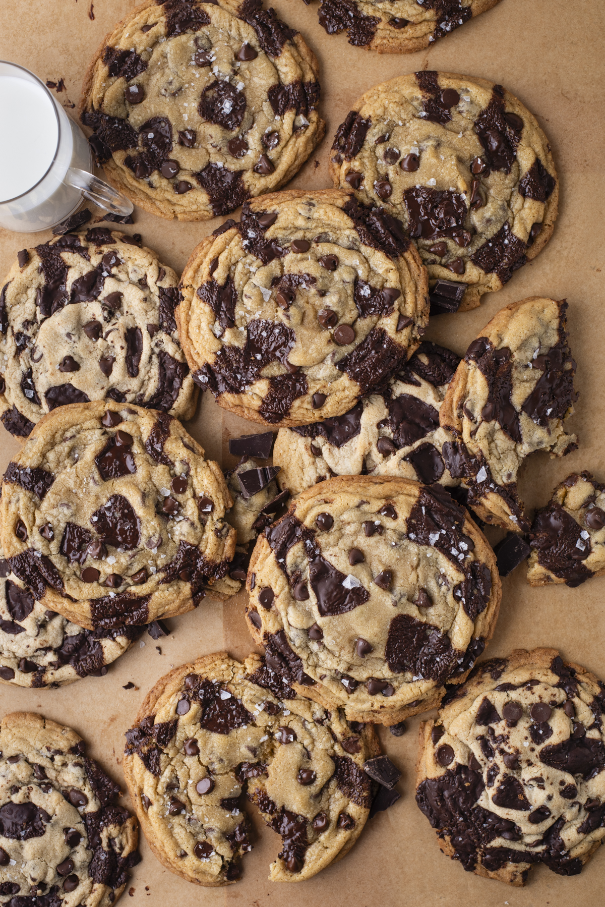 A group of large chocolate chip cookies with chunks of dark chocolate and a sprinkling of sea salt, arranged on brown parchment paper next to a glass of milk.
