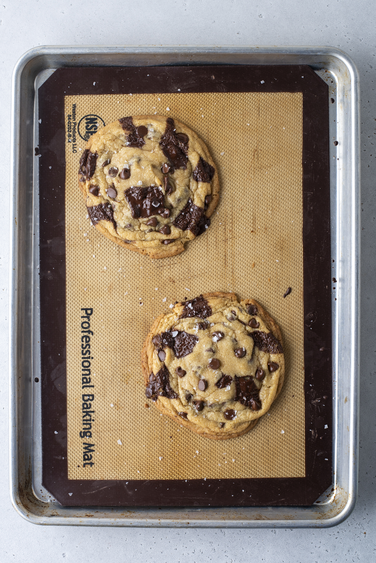 A baking sheet with a silicone mat holds two large chocolate chip cookies sprinkled with sea salt. The cookies are golden brown with chunks and chips of chocolate, resting on a light grey countertop.