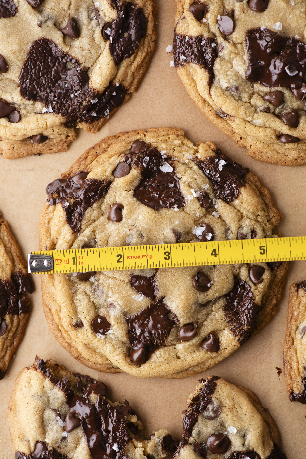 A close-up of large chocolate chip cookies on parchment paper, with a yellow measuring tape across one cookie showing it is about 4 inches wide. The cookies have visible chocolate chunks and chips.