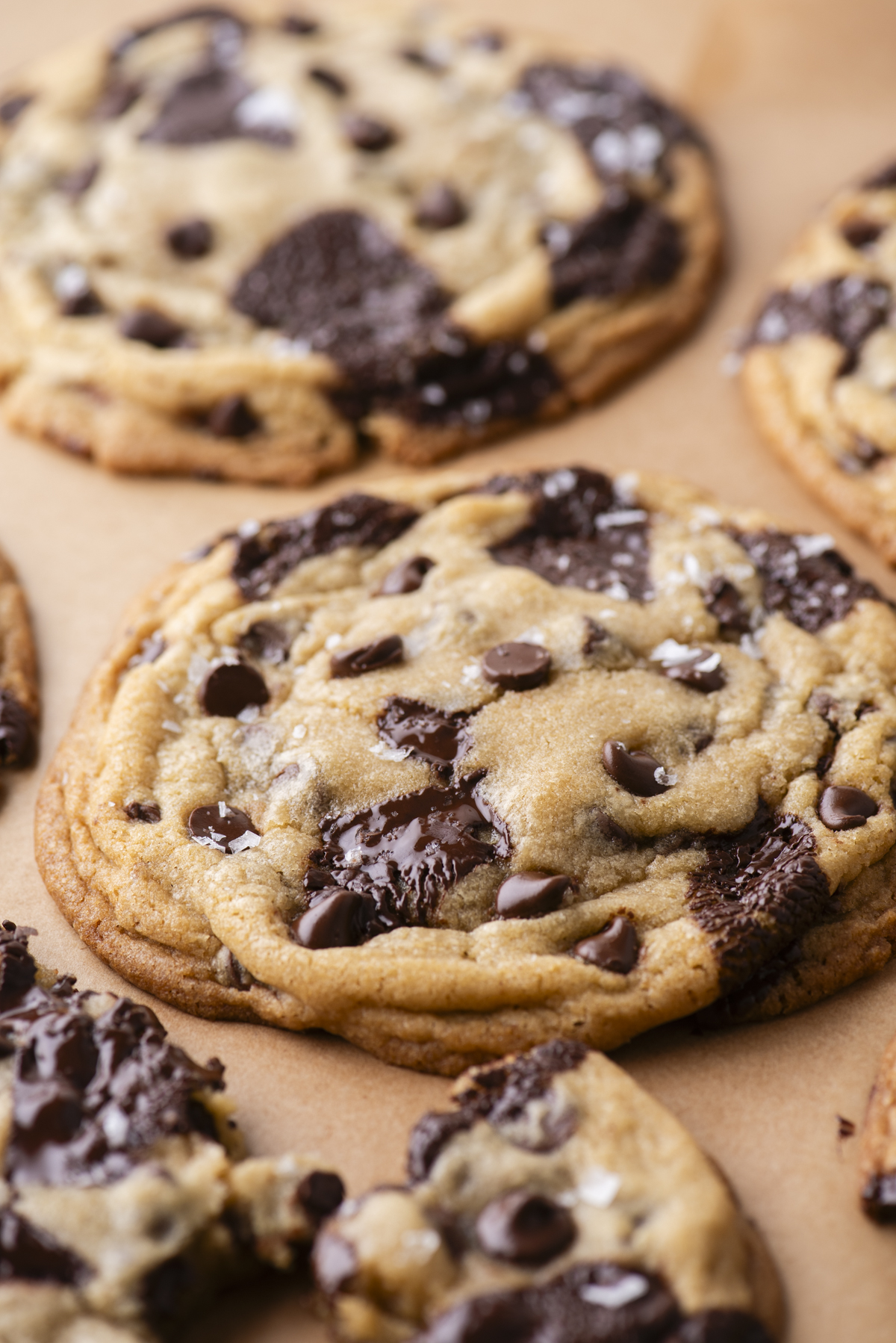 Close-up of freshly baked chocolate chip cookies with melted chocolate chunks on brown parchment paper. The cookies are golden brown and look soft and chewy.