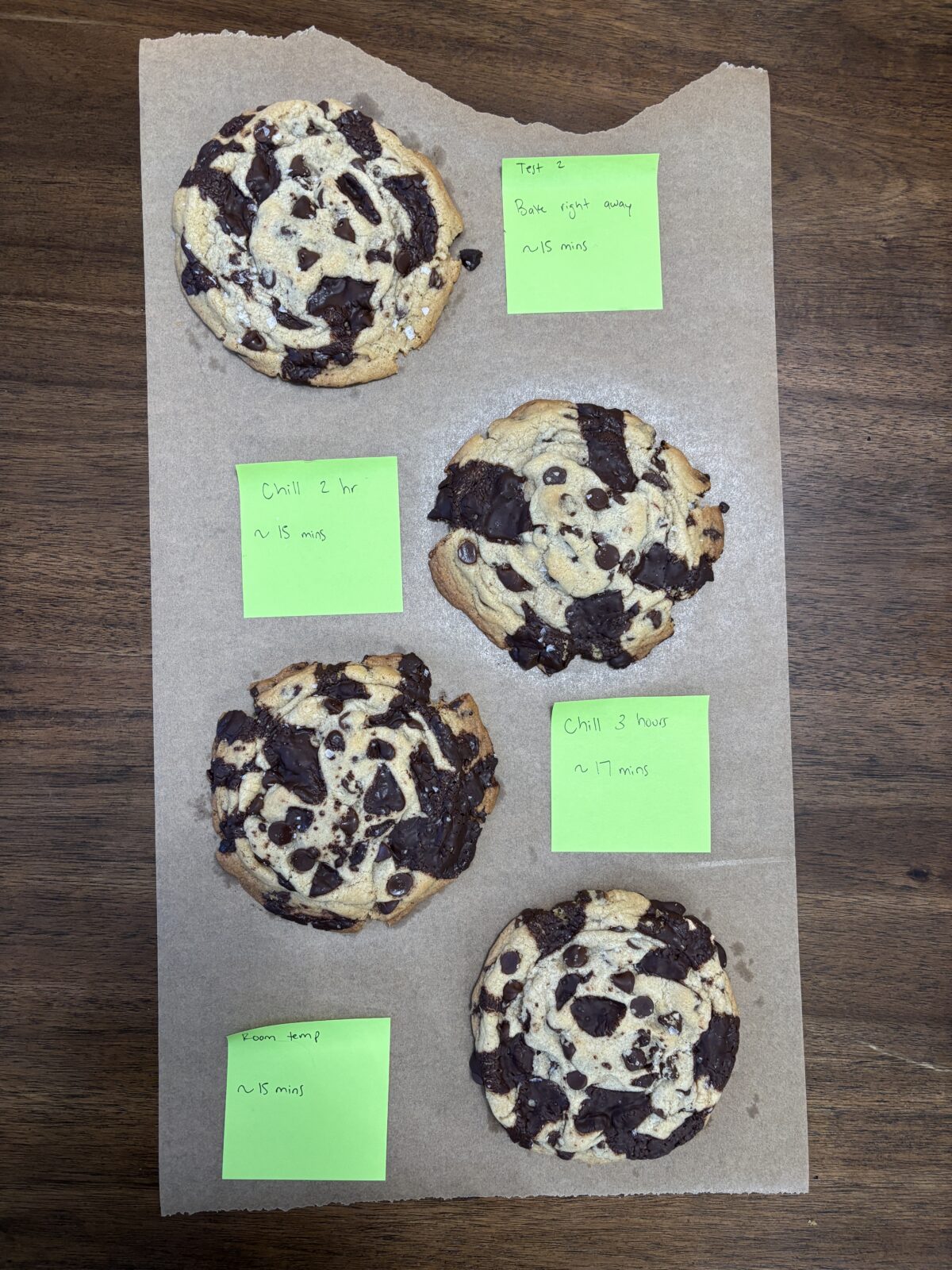 Four chocolate chip cookies on parchment paper, each labeled with green sticky notes describing different chilling times and baking durations for recipe testing, on a wooden surface.