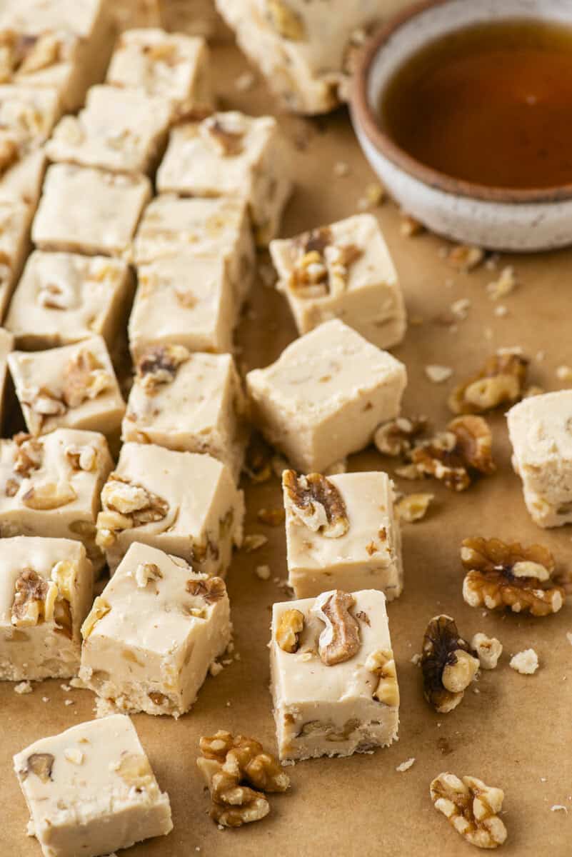 Cubes of maple walnut fudge are arranged on brown parchment paper, surrounded by scattered walnut pieces. A small bowl of maple syrup sits nearby.