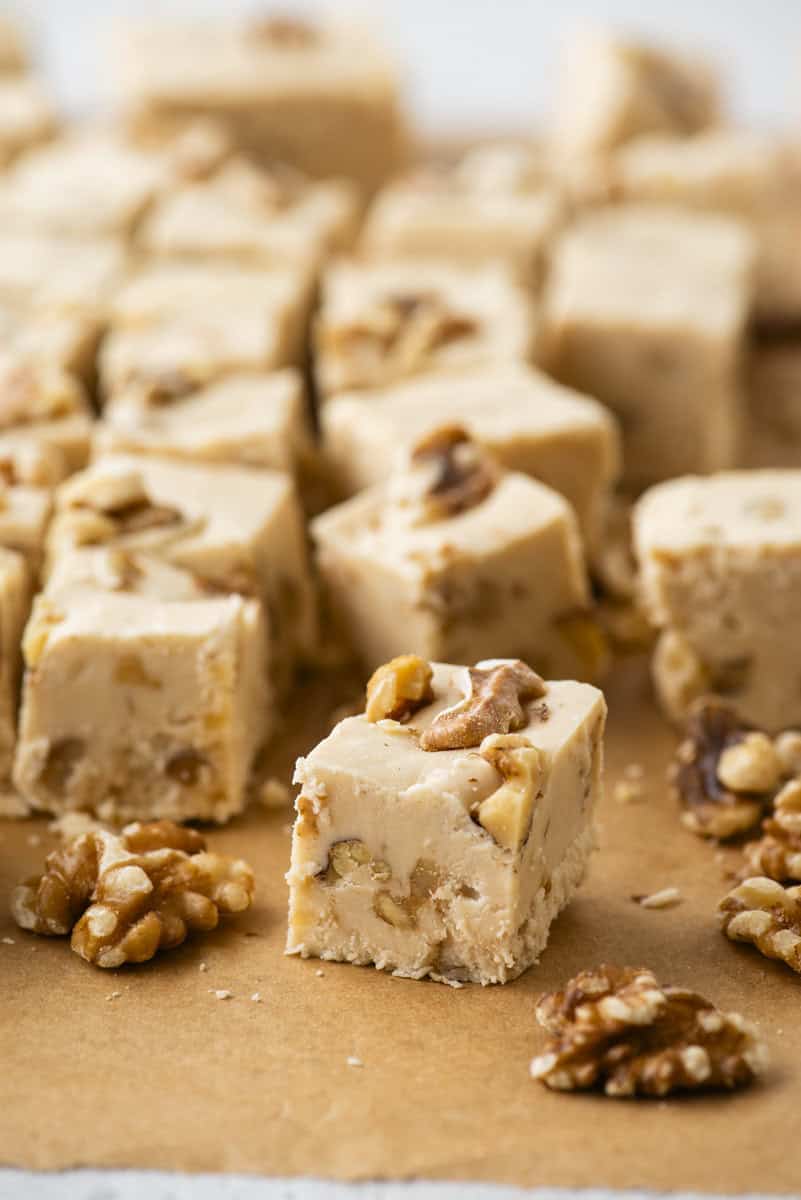 Close-up of creamy fudge squares with pieces of walnuts on top and inside, arranged on brown parchment paper. Some walnut halves are scattered around the fudge pieces.