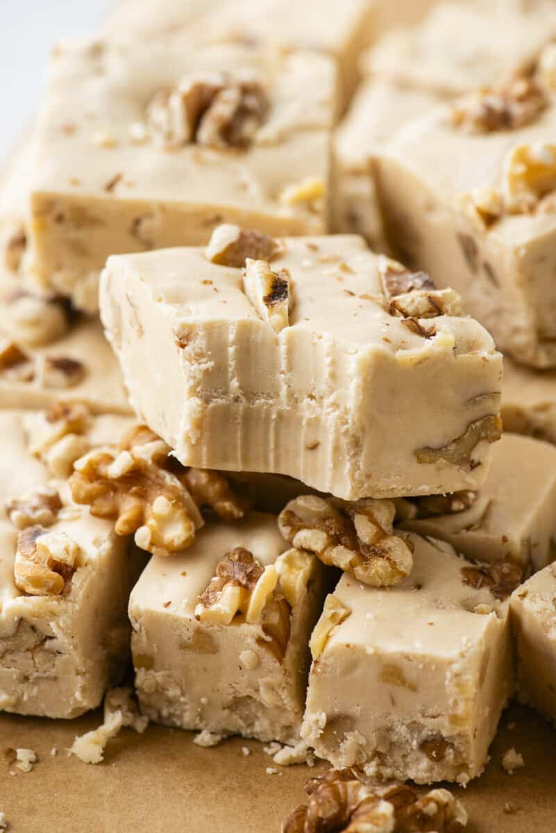 Close-up of creamy fudge squares with visible walnut pieces, stacked together on brown parchment paper. Some pieces show bite marks, highlighting the smooth, dense texture and crunchy nuts.