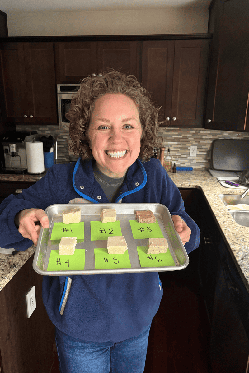 A smiling person with curly hair holds a baking tray with six pieces of dessert, each labeled with a number on green paper, standing in a kitchen with dark cabinets and a granite countertop.