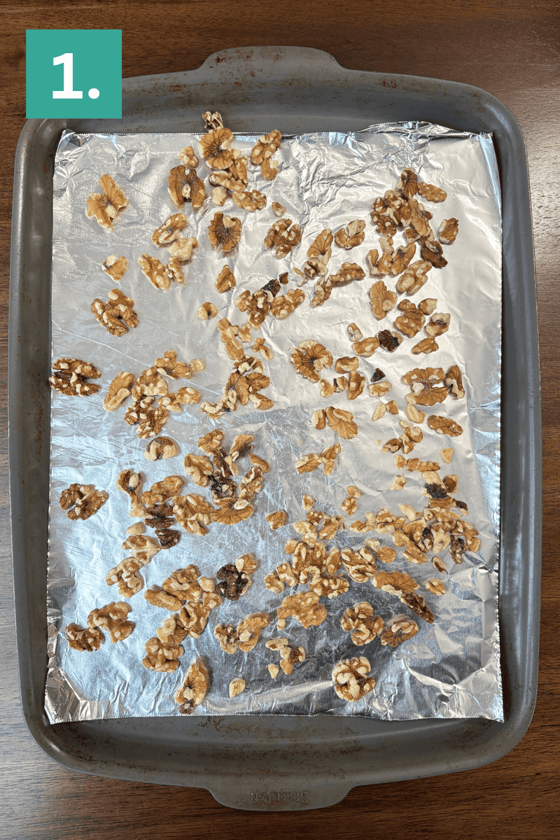 A baking tray lined with aluminum foil holds scattered walnut pieces, ready to be roasted. A green box with the number 1 appears in the top left corner. The tray sits on a dark wooden surface.