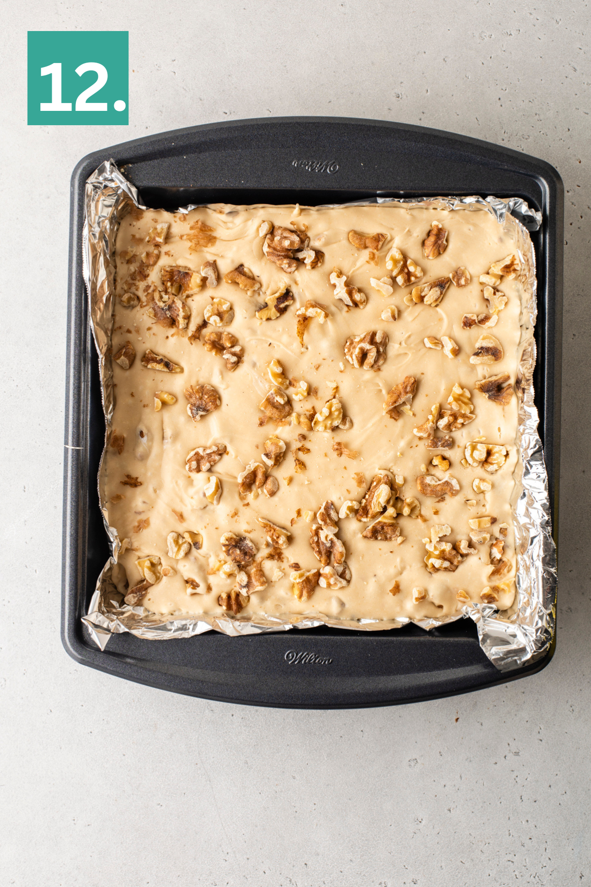 A square baking pan lined with foil holds a layer of beige fudge topped with chopped walnuts. A green box with the number 12 is in the top left corner. The pan rests on a light gray surface.