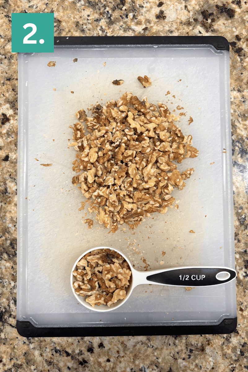 Chopped walnuts on a white cutting board, with a 1/2 cup measuring cup filled with walnuts. A green box with the number 2. is in the top left corner. The cutting board is on a granite countertop.