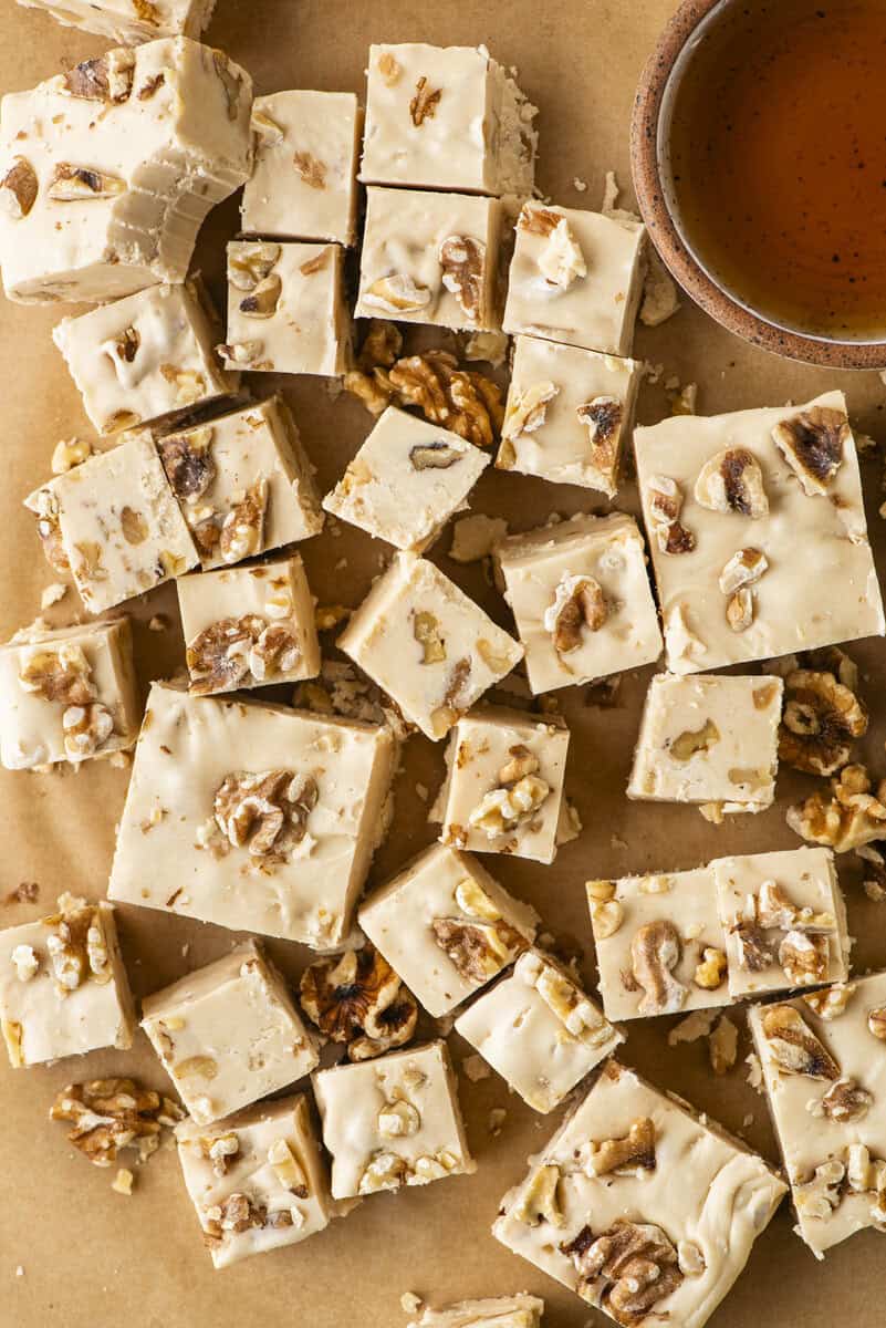 Squares of creamy maple walnut fudge topped with chopped walnuts are scattered on brown parchment paper next to a small cup of maple syrup.