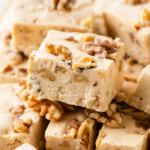 Close-up of creamy fudge squares with pieces of walnuts mixed in and scattered on top, stacked closely together.