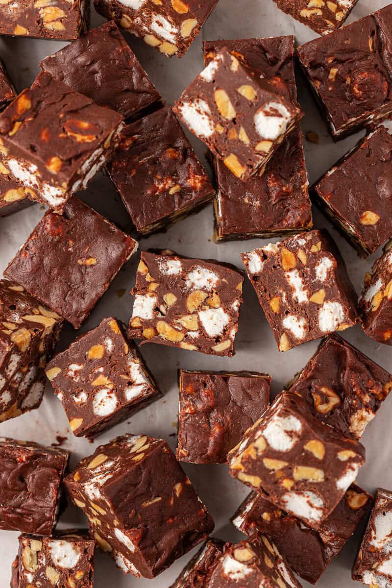 Squares of chocolate fudge filled with chopped nuts and mini marshmallows are arranged closely together on a light surface, showing the mix of ingredients inside each piece.