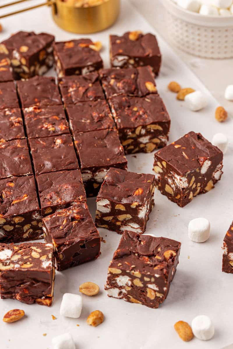 Squares of chocolate fudge with visible peanuts and mini marshmallows are arranged on parchment paper, with some pieces separated. Loose peanuts and mini marshmallows are scattered around.