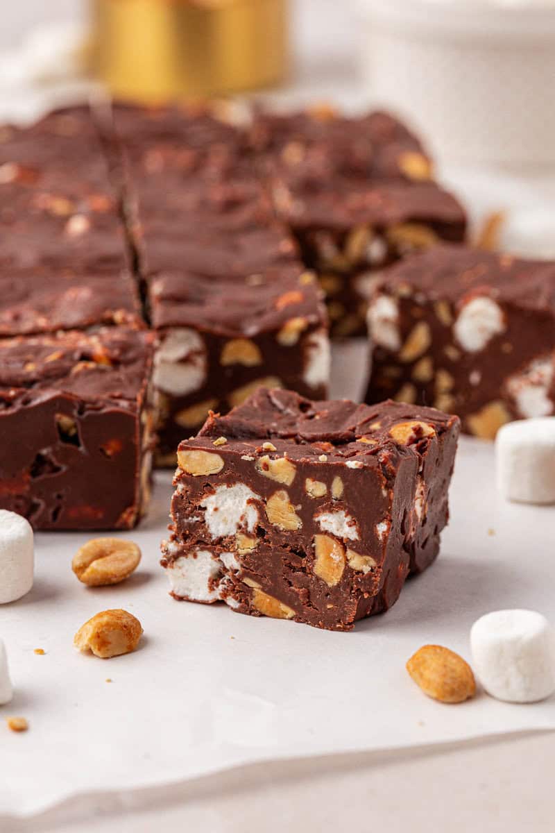 A close-up of chocolate fudge squares with visible peanuts and mini marshmallows inside, surrounded by a few loose peanuts and marshmallows on a white surface.