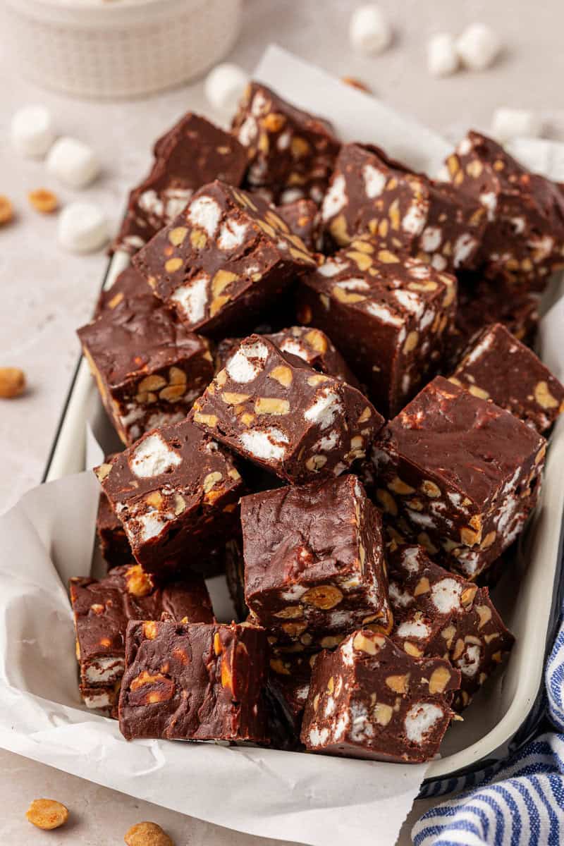 A tray filled with squares of rocky road fudge, showing chunks of marshmallows and peanuts inside the rich chocolate. Some peanuts and mini marshmallows are scattered around the tray.