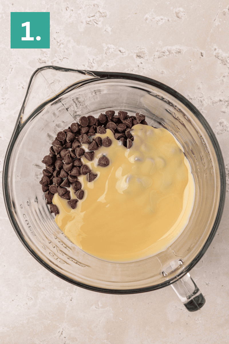 A glass mixing bowl containing chocolate chips and sweetened condensed milk sits on a light countertop. A green box with the number 1 is in the upper left corner, indicating the first step of a recipe.