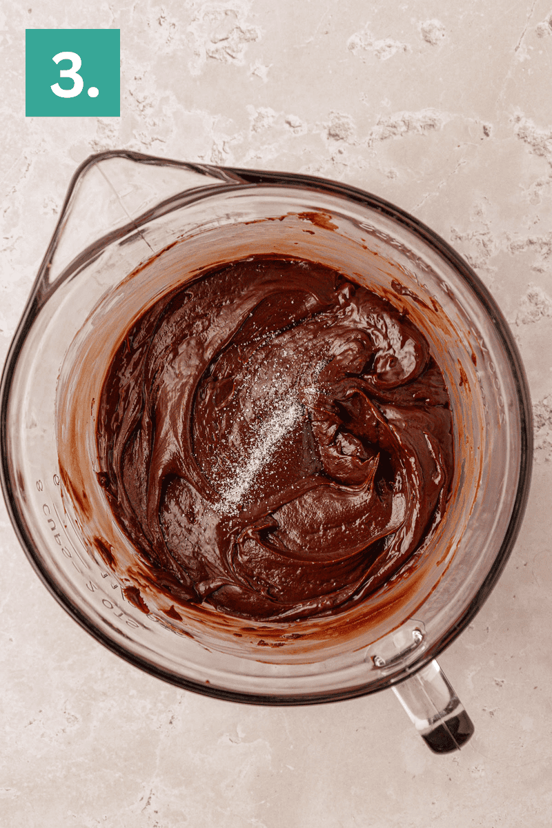 A glass mixing bowl filled with thick chocolate batter and a sprinkle of salt on top, placed on a light beige surface. A green box with the number 3 is in the upper left corner.