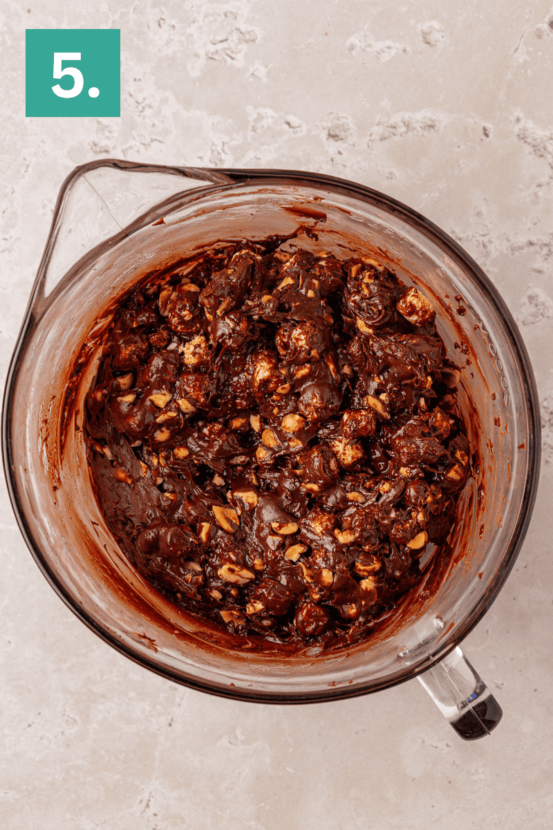 A glass mixing bowl filled with chocolate batter mixed with nuts and marshmallows sits on a light countertop. A green box with the number 5 is in the top left corner.