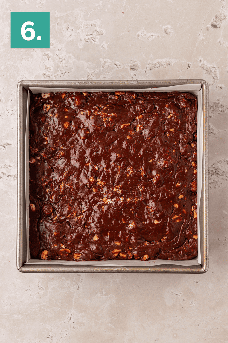 A square metal baking pan filled with unbaked chocolate brownie batter, resting on a light-colored, textured countertop. A green square with the number 6 is in the top left corner.