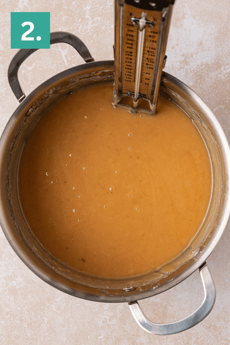 A saucepan filled with light brown caramel mixture sits on a countertop, with a candy thermometer clipped to the side. A green box in the corner displays the number 2.