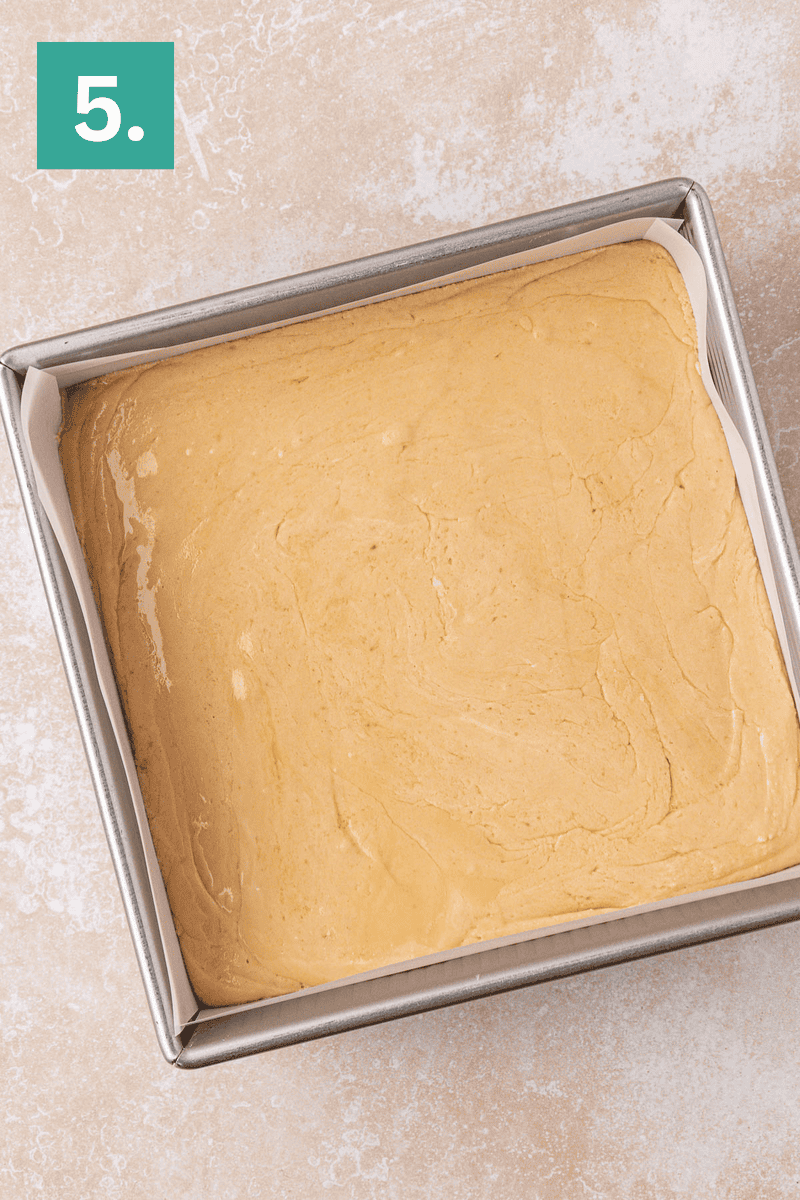A square baking pan lined with parchment paper, filled with smooth, light brown batter, sits on a light countertop. A green box with the number 5 is in the top left corner.