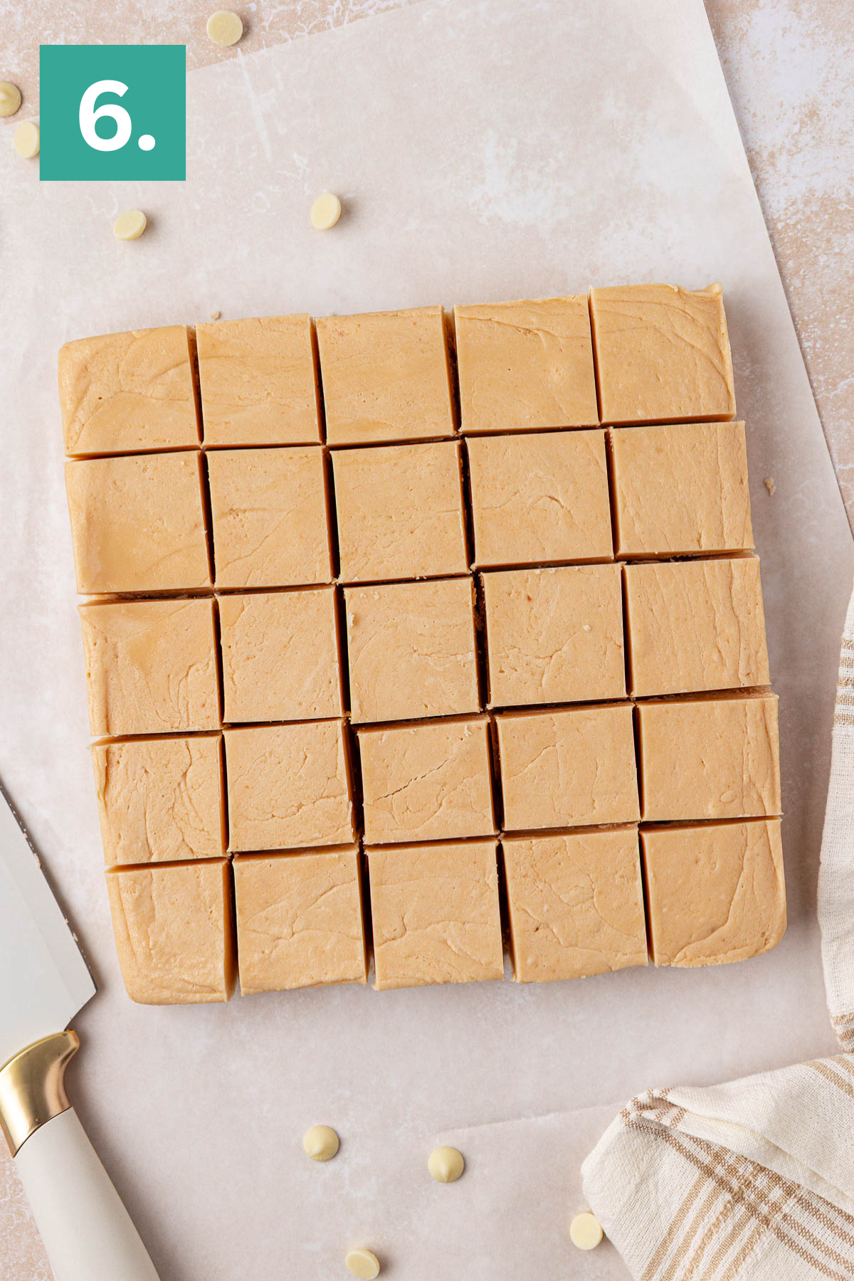 A square block of light brown fudge cut into 25 even pieces sits on parchment paper. A knife with a white handle and a beige-striped cloth are beside it. A green square with the number 6 is in the top left corner.