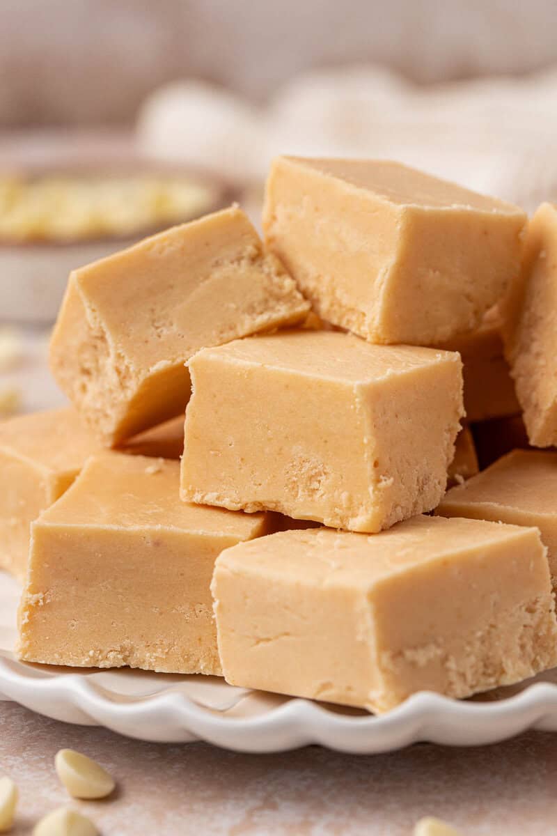 A plate stacked with creamy, light brown peanut butter fudge squares, with a smooth texture and a few white chocolate chips scattered around on the surface.