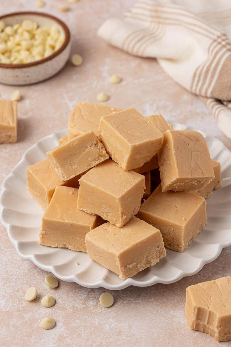 A plate stacked with creamy beige fudge squares, surrounded by scattered white chocolate chips. A bowl of more chocolate chips and a striped cloth are in the background.