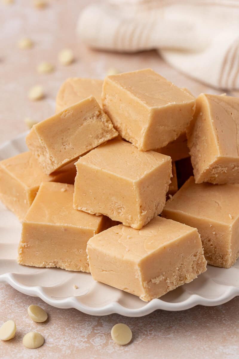 A stack of creamy, light brown fudge squares arranged on a white, scalloped-edged plate with a few white chocolate chips scattered nearby.