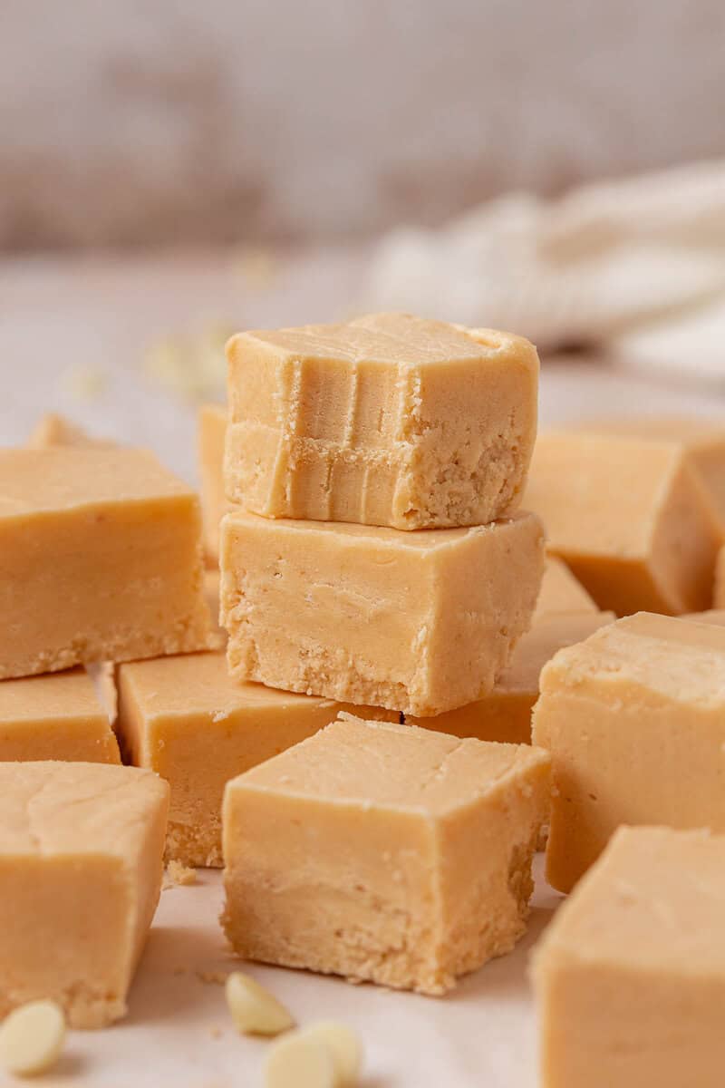 A stack of creamy, light brown fudge squares sits on a surface, with the top piece showing bite marks. More fudge pieces and white chocolate chips are scattered around.