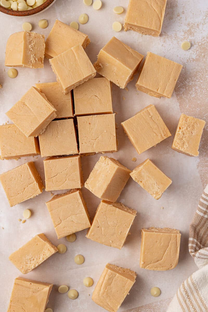 Squares of creamy peanut butter fudge are arranged on parchment paper, with a few white chocolate chips scattered around. A striped cloth is partially visible in the corner.