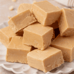 A stack of creamy, light brown fudge squares arranged on a white, scalloped plate. The fudge appears smooth and slightly crumbly at the edges.